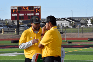 BLPA football coaches Matt Pacheco and Josh Pacheco talking on football field with scoreboard in the distance.